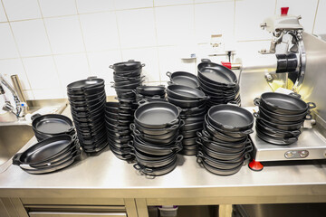 Large stack of clean black serving pans organized on a stainless steel kitchen counter in a...