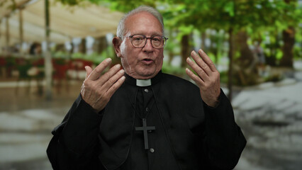 Senior priest man wearing glasses gestures outdoors on a street, with blurred green trees and...