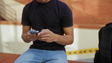 Young man outdoors using smartphone on city street sitting on brick wall, dressed casually in black shirt and jeans, surrounded by urban setting with focus on device interaction.