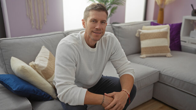 Young man sitting on a comfortable gray sofa in a stylish living room with soft pillows and pastel walls, exuding a relaxed and inviting atmosphere indoors.