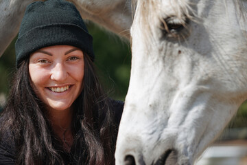 Young woman in beanie standing near her white Arabian horse on ranch farm, smiling