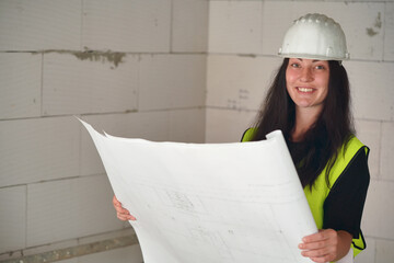 Young female construction worker in green high visibility vest and white hard hat, holding paper drawings with building documentation blueprint, smiling to camera, blurred bare walls background