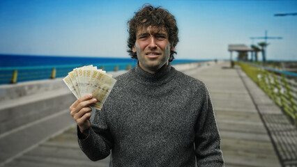 Hispanic man holding norwegian krone banknotes stands on a seaside pier enjoying the outdoor beach view with a casual sweater in clear sunny weather.