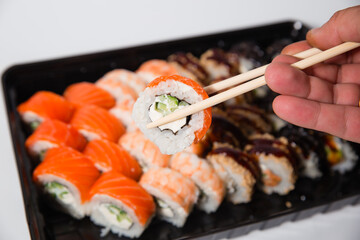 Delicious dinner, on the table various Sushi rolls with salmon, tuna, smoked eel, avocado, cream cheese laid out in a box. Sushi menu. Top view of various sushi. Holding sushi with chopsticks.