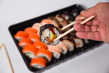 Delicious dinner, on the table various Sushi rolls with salmon, tuna, smoked eel, avocado, cream cheese laid out in a box. Sushi menu. Top view of various sushi. Holding sushi with chopsticks.