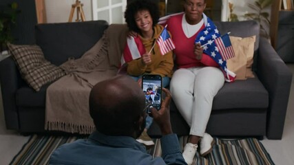 Black man using smartphone to take pictures of gen alpha son and wife posing for camera with American flags on couch on Fourth of July at home - Powered by Adobe