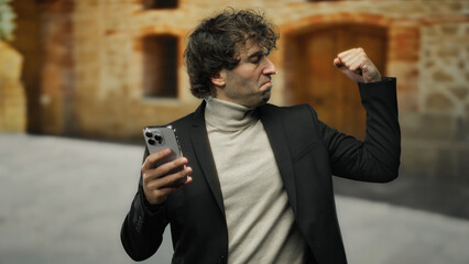 Young man makes a confident gesture while holding a smartphone in an urban outdoor setting, dressed...