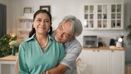 Fototapeta premium Happy senior couple with casual cloth spending time together and hugging at modern kitchen. Grandmother and grandfather holding hand together and looking at camera. Healthy relationship. Myrmidon.