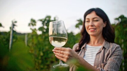 Happy Caucasian woman smiling with joy while looking at glass of white wine. Standing before bushes of grapes. Enjoying taste of fresh drink. Winemaking concept. Observing purity of alcohol.