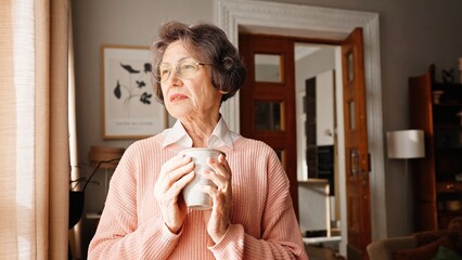 Excited old woman standing by window and looking outside at street. Sad grandmother holding cup of water or hot drink. Feeling lonely. Expecting visit from her family. Old age concept.