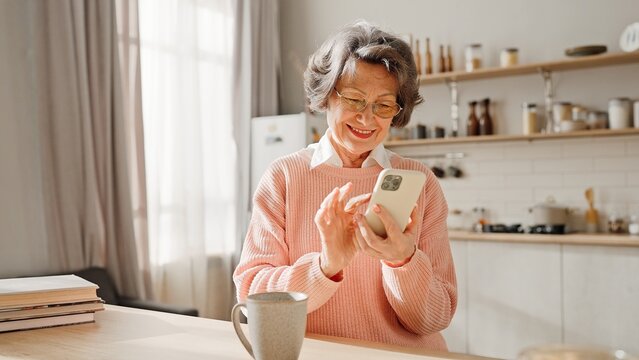 Good-looking mature Caucasian woman with casual clothes spending time in cosy kitchen. Happy grandmother watching positive video or family photo on cell phone. On table standing cup or hot drink.