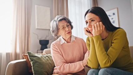 Two women sitting on couch in living room. Elderly female in pink sweater talking with daughter....