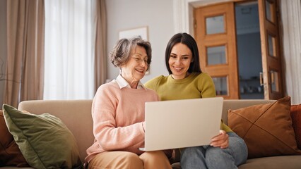 Positive Caucasian grandmother with her daughter or granddaughter using laptop. Women talking by...