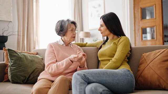 Two Caucasian people hugging each other while sitting on sofa. Mother and daughter looking happy. Smiling to each other while expressing love and care. Cosy flat interior in background.