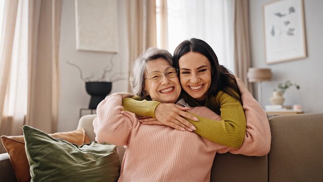 Elderly Caucasian woman with grey hair wearing glasses while reading book. Charming daughter walking up behind mature mother and hugging her. Older mom widely smiling while looking at her relative.
