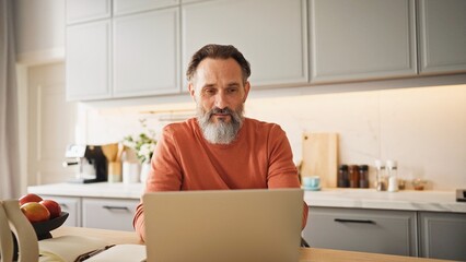 Handsome bearded man keeping his hand on chin while watching at laptop screen. Adult male moving his head from side to side and smiling with joy. Checking his mail during breakfast in kitchen.