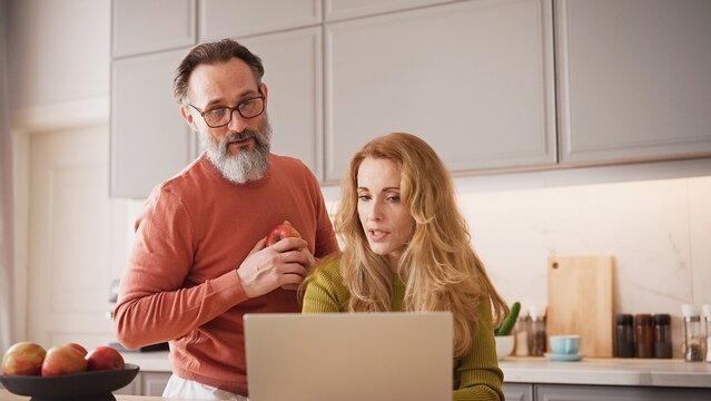 Lovely Caucasian couple spending time together in middle of modern kitchen. Woman using laptop while her husband with beard and glasses stands behind her. Man holding apple. Discussing purchase. - Powered by Adobe