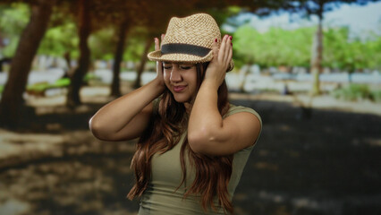 Hispanic woman in park wearing hat covers ears due to noise amid green trees and shadows outdoors.