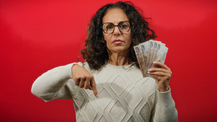 Woman in glasses pointing down while holding russian rubles against a vibrant red background, appearing serious and focused, wearing a knitted sweater.