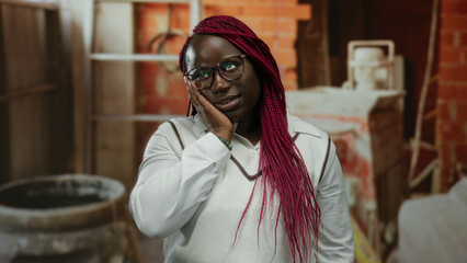 Woman with glasses and red braids stands at indoor construction site displaying thoughtful expression and confident demeanor.