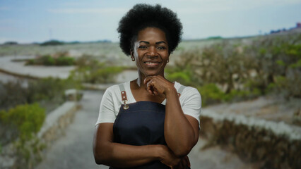 Confident woman in apron stands outdoors in a park setting, exuding warmth and friendliness,...