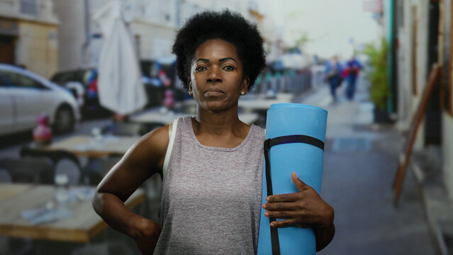 Woman holding yoga mat stands on outdoor terrace of a vibrant city street restaurant, embodying urban tranquility. - Powered by Adobe
