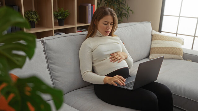 Pregnant woman sitting on a sofa in a cozy living room using a laptop, illustrating a relaxed indoor atmosphere with a modern interior and home comfort.