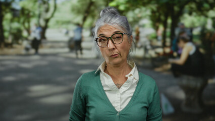 Fototapeta premium Senior woman with grey hair crossing arms seriously in a outdoor park setting surrounded by lush greenery and people in the background.