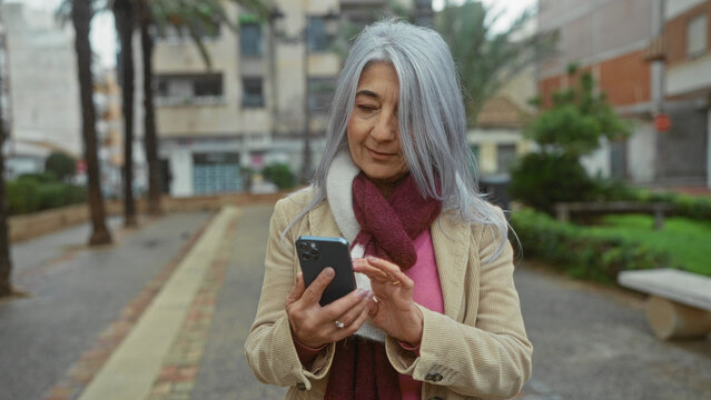 Senior woman with grey hair using smartphone outdoors on an urban street, wearing a scarf and coat in a calm and modern environment with palm trees. - Powered by Adobe