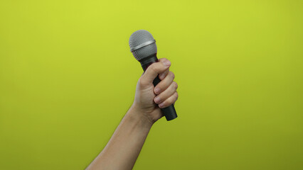 Man holding microphone against vibrant yellow wall, showcasing male hand gripping mic with green...