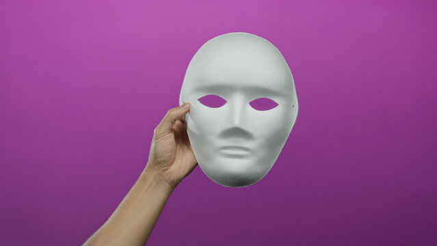 Man holding a blank white mask against a vibrant pink background, showcasing elements of anonymity and creativity isolated in a studio shot.