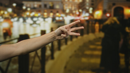 Man showing three fingers on a street with blurred urban lights, highlighting human presence amid city life during evening, emphasizing hand gesture in an outdoor setting.
