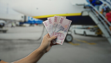 Man holding turkish lira banknotes outdoors at an airport near a plane in terminal 1, capturing travel and finance themes in a dynamic setting. © Krakenimages.com