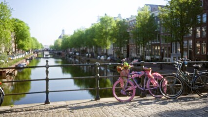 Blurred bicycles on a bridge with amsterdam canal and trees in the out-of-focus background, creating a serene bokeh effect under a clear sky.