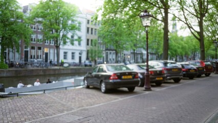 Blurred view of amsterdam canal with parked cars and people passing by in a serene urban street scene enveloped in green foliage and traditional architecture.