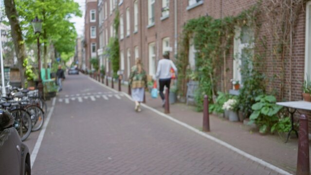 Defocused view of a european city street with a man and woman walking along a brick path lined with greenery, showcasing urban life in a tranquil town setting.