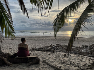 Frau sitzt unter Palmen an einem Strand bei Puntarenas auf der Halbinsel Nicoya und blickt auf den Sonnenuntergang, Costa Rica
