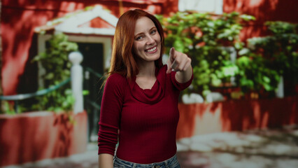 Woman standing on city street smiling and pointing with red hair wearing red top in urban outdoor setting, with plants and buildings in the background.