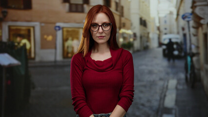 Obraz premium Woman with red hair wearing glasses and sweater standing on city street with historical buildings in background, conveying urban lifestyle and elegance.