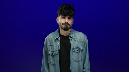 Young man with denim jacket posing isolated over blue background looking contemplative