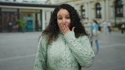 Young hispanic woman in green sweater outdoors on bustling street, expressing surprise with hand over mouth, city background adds urban context and modern feel.