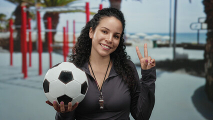 Woman smiling holding soccer ball outdoors near gym with beach background wearing black, displaying...