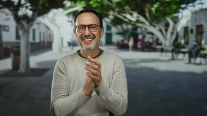 Man smiling and clapping in an urban park setting, surrounded by trees and city architecture,...