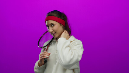 Woman posing with tennis racket on a vibrant pink background, exuding youthful energy and showcasing her athletic attire and playful demeanor while touching her cheek.