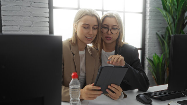 Two blonde women executives collaborate in a modern office setting with digital tablet in hand, illustrating teamwork and innovation in a professional environment.