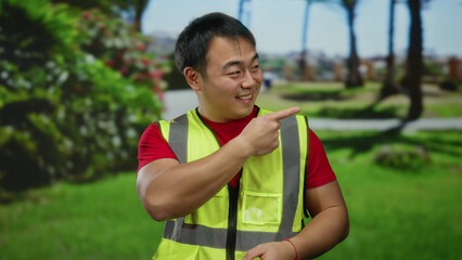 Young man in a green park wearing a safety vest smiling and pointing with both hands showcasing various directions and gestures with a blurred natural background.