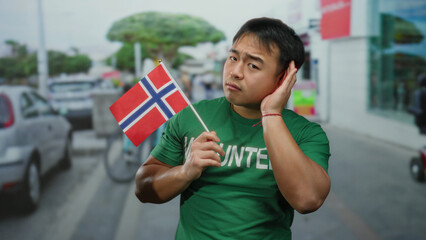 Young man holding a norwegian flag gestures listening on a busy urban street with blurred...