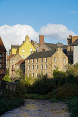 Naklejka premium A sunny afternoon in Edinburgh’s Dean Village, where charming stone houses bask in the golden light, framed by a clear blue sky and fluffy white clouds above the peaceful riverside setting.