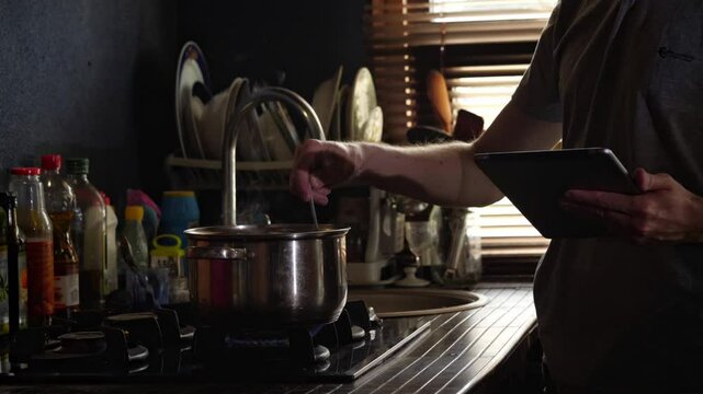 A mid-shot captures a focused and modern mood as a man stirs a pot on a gas stove while referencing a digital tablet, suggesting technology-assisted home cooking in a dimly lit kitchen