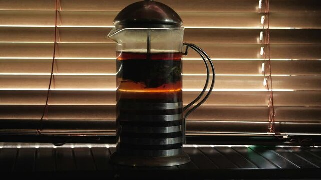 A close-up shot captures a warm, intimate mood as a French press, filled with brewing tea or coffee, is silhouetted against the soft light filtering through horizontal window blinds